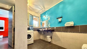 A modern bathroom with a blue accent wall, a washing machine, a sink with a towel rack, and a toilet. - "  Happy  Apartment " (Bari Sardo)
