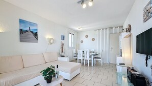 Ein modernes Wohnzimmer mit einer Couch, einem kleinen runden Tisch mit einer Pflanze, einem Essbereich mit weißem Tisch und Stühlen sowie einem an der Wand befestigten Fernseher.