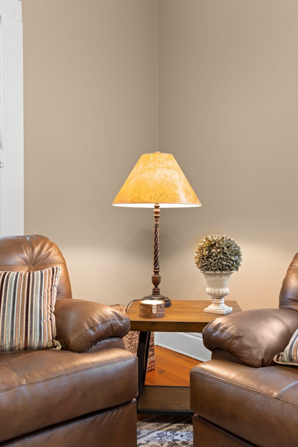 Een hoek van een kamer met twee bruine leren stoelen, een klein houten bijzettafeltje met een lamp en een decoratieve plant, en een witte deur op de achtergrond.