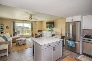 Una cocina moderna con una isla central, un comedor con sofá, un televisor montado en la pared y un balcón con vistas.