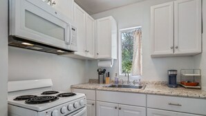 Une cuisine avec des meubles blancs, une cuisinière, un four à micro-ondes, un évier et une fenêtre avec un rideau.