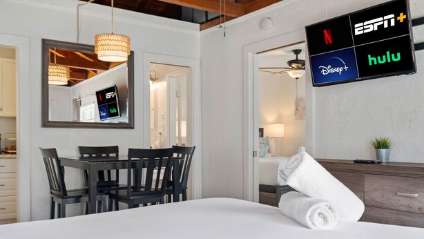 Une chambre d’hôtel comprenant un lit, un téléviseur affichant les logos de services de diffusion en continu, un coin repas avec une table et des chaises, et une salle de bain avec une serviette posée sur le comptoir.