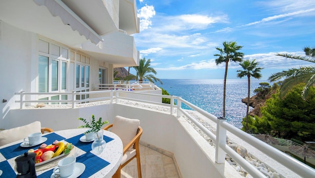 A balcony with a table set for breakfast, overlooking a scenic ocean view with palm trees and rocky terrain. - Panorama by Interhome (Altea)