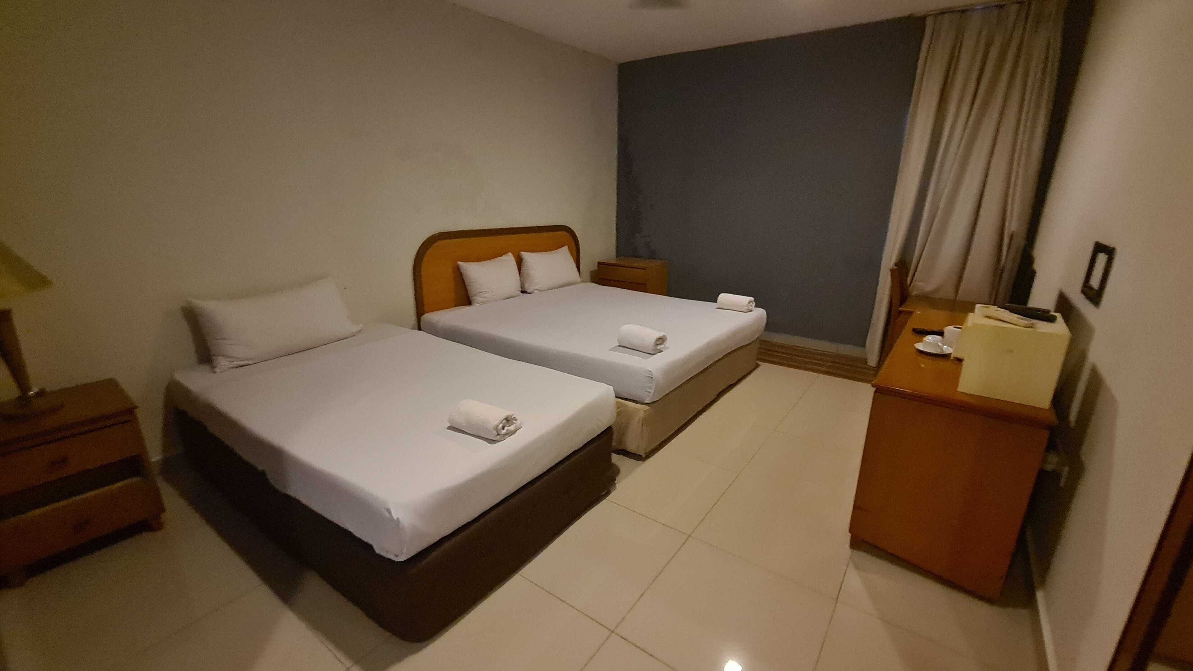 A hotel room with two single beds, a wooden nightstand, a desk with a chair, and a small table with a lamp.