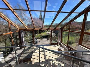 A glass-enclosed outdoor area with a wooden deck, seating, and a view of a garden.