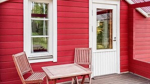 Een rood houten gebouw met een witte deur en ramen, een houten terras en twee houten stoelen en een tafel.