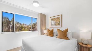 A hotel room with a large bed, a ceiling fan, a window with a view of palm trees, and a framed picture on the wall.