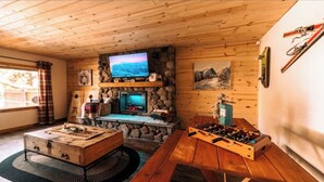 A cozy cabin interior with a stone fireplace, a television mounted on the wall, a wooden table with a foosball table, and a rustic wooden chest.