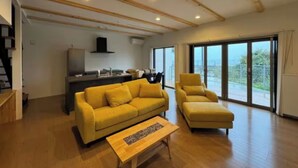 Ein modernes Wohnzimmer mit einem gelben Sofa, dazu passendem Hocker und einem Couchtisch aus Holz. Im Hintergrund ist eine Küchenzeile erkennbar, über dem Herd hängt eine schwarze Dunstabzugshaube.