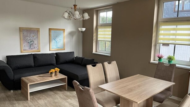 A modern living room with a black sofa, a wooden coffee table, a dining table with chairs, and two framed artworks on the wall. - Vacation apartment Lehesten Zinkenweg (Lehesten)