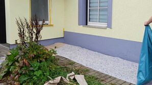 A house with a yellow exterior and a grey roof. There is a window with a blue frame and a window blind. A person is holding a blue bag. - Vacation apartment Lehesten Zinkenweg (Lehesten)