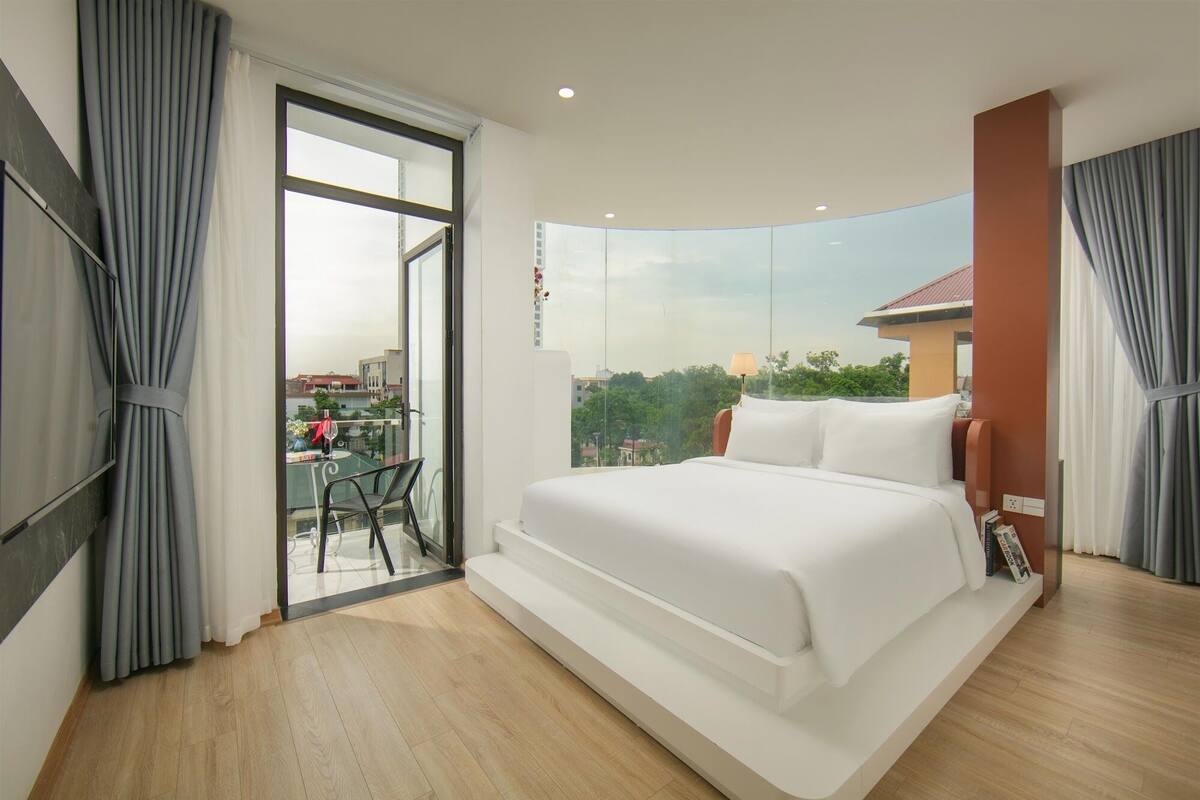 Une chambre moderne avec un grand lit, un balcon doté d’une table à manger et une vue sur la ville.