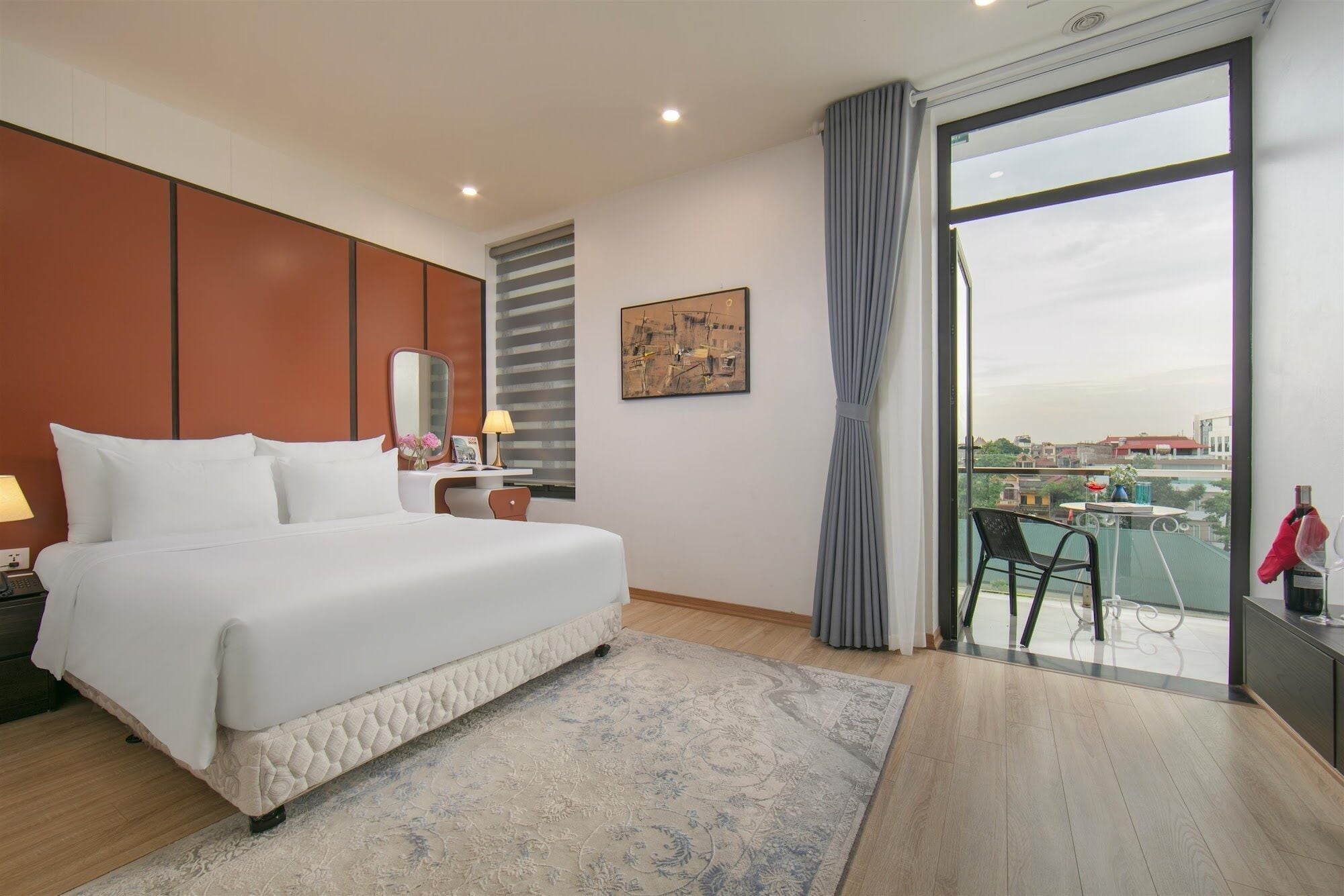 Une chambre d’hôtel avec un grand lit, un bureau avec une lampe, un tableau au mur et un balcon offrant une vue sur la ville.