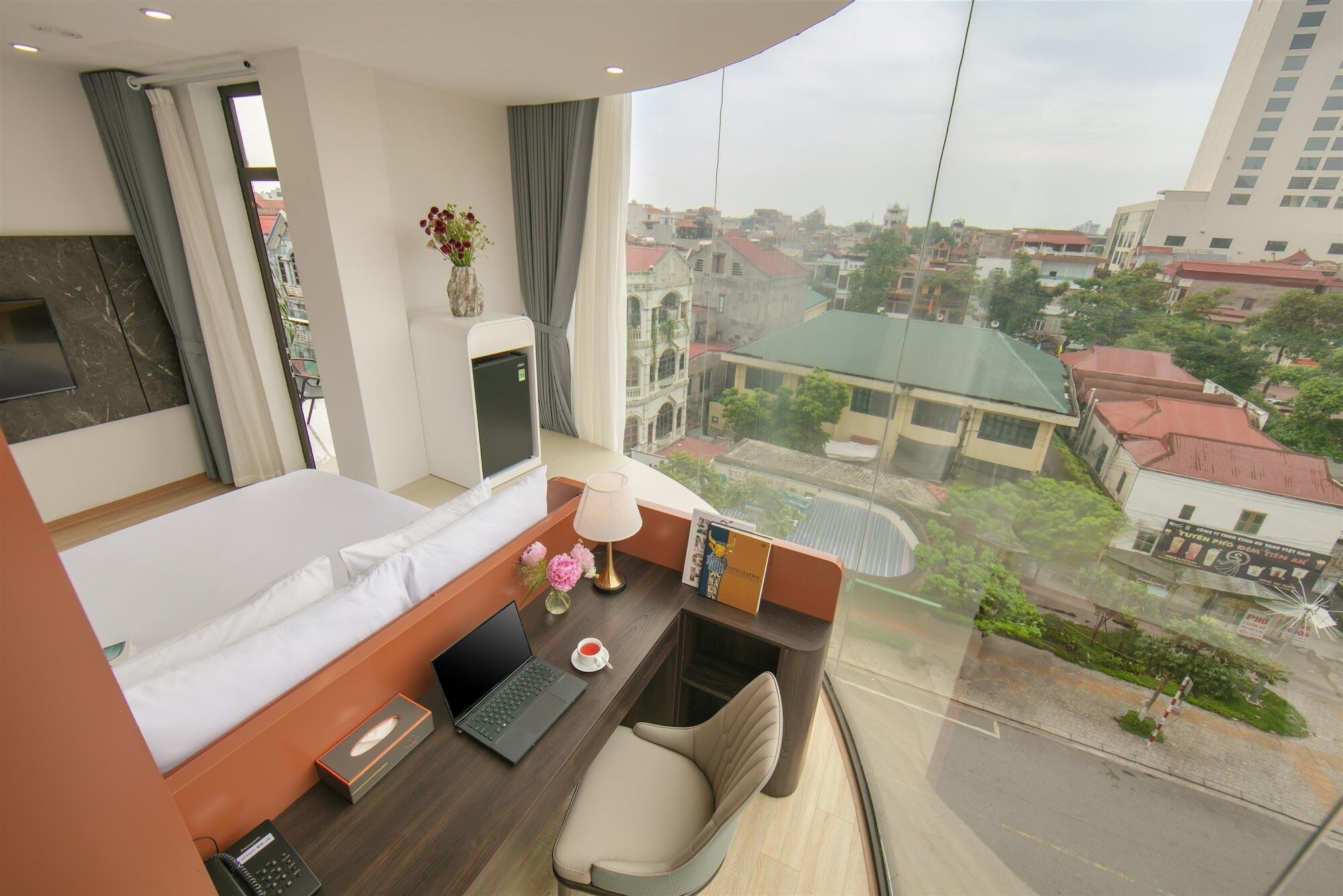 Une chambre d’hôtel avec un lit, un bureau avec un ordinateur portable, une chaise, une petite table avec une lampe et une vue sur le paysage urbain.