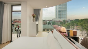Una habitación de hotel moderna con una cama grande, una mesita con lámpara, un jarrón con flores y vistas a un paisaje urbano con edificios y árboles.