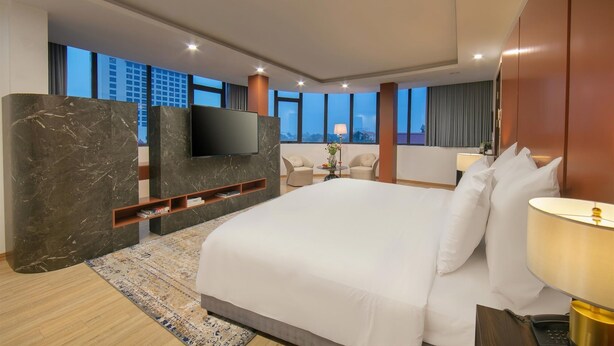 Una habitación de hotel moderna con una cama grande, un televisor de pantalla plana montado en una pared de mármol, una pequeña zona de estar con dos sillones y vistas a la ciudad a través de grandes ventanales.