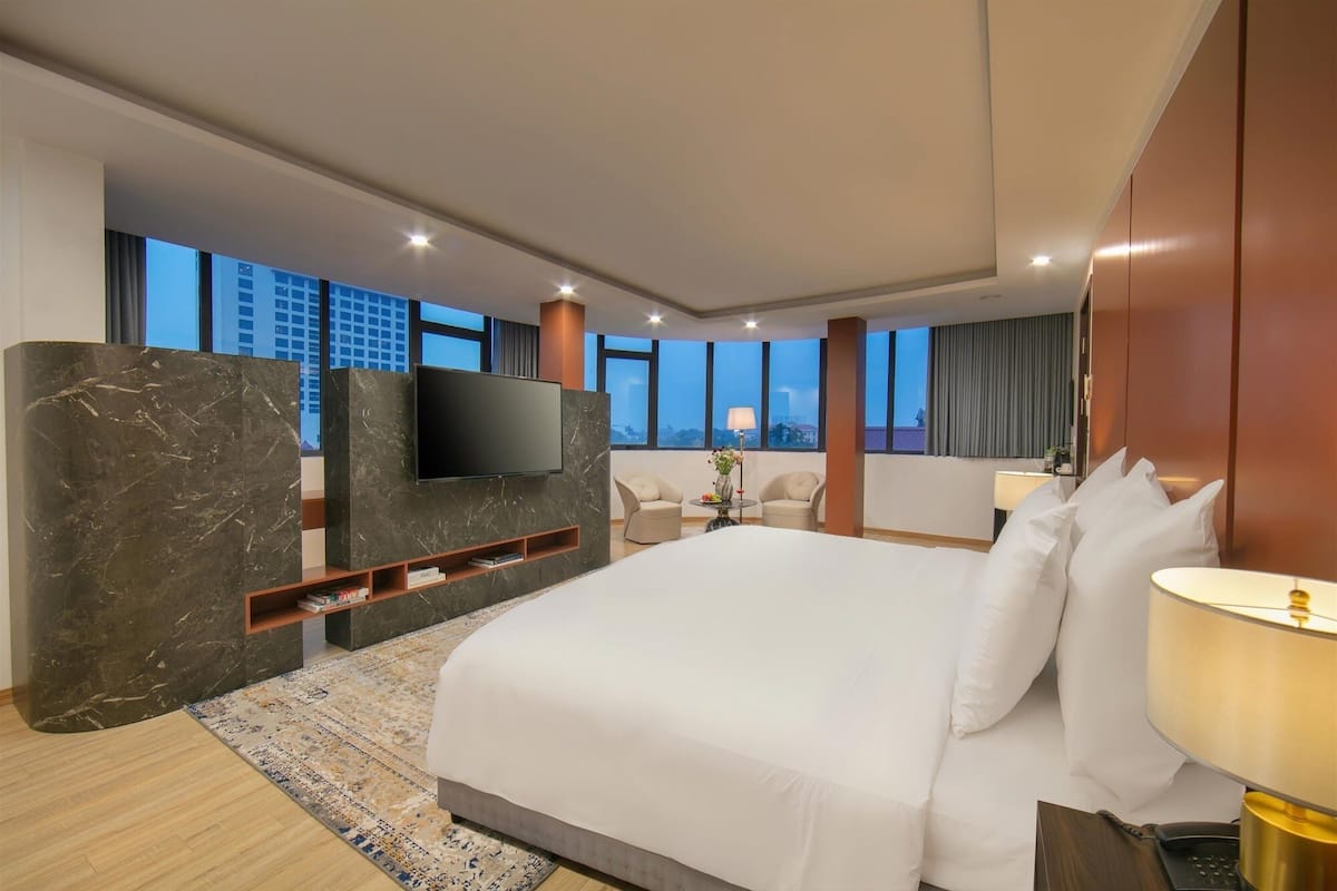 Une chambre d’hôtel moderne dotée d’un grand lit, d’un téléviseur à écran plat fixé sur un mur en marbre, d’un petit coin salon avec deux fauteuils et offrant une vue sur le paysage urbain grâce à de grandes fenêtres.