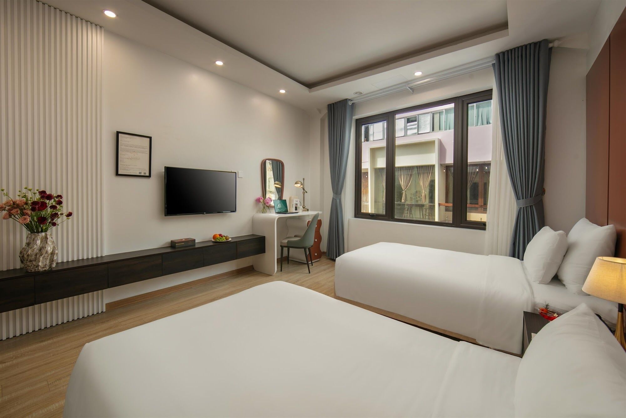 Habitación de hotel con dos camas, un escritorio con silla, televisor de pantalla plana, ventana grande con cortinas y un florero con flores.