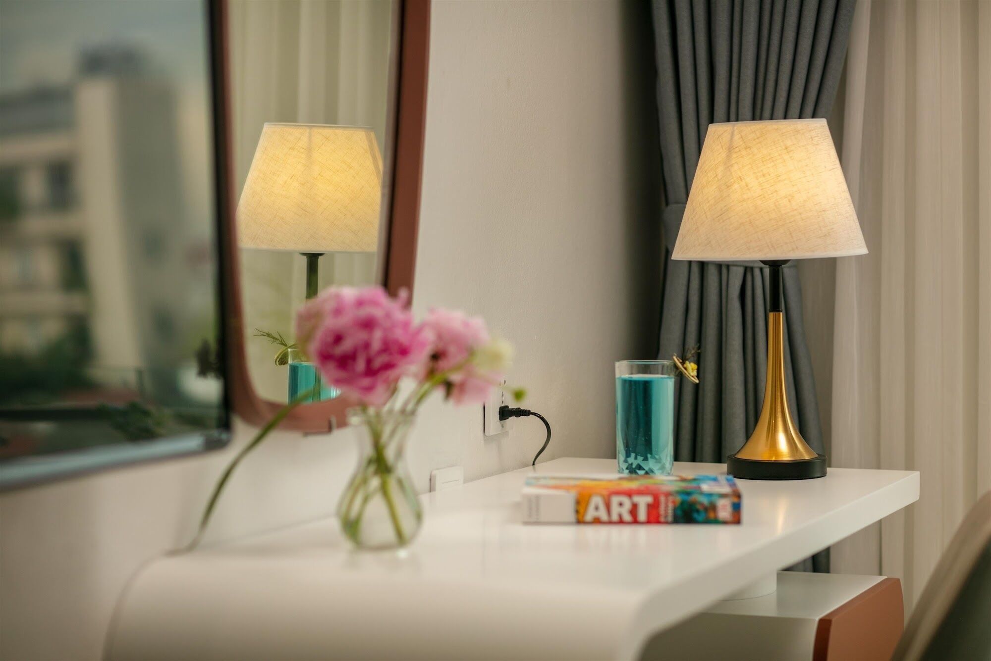 A hotel room with a white console table, two lamps, a vase with pink flowers, a glass of blue liquid, and a book titled "ART".