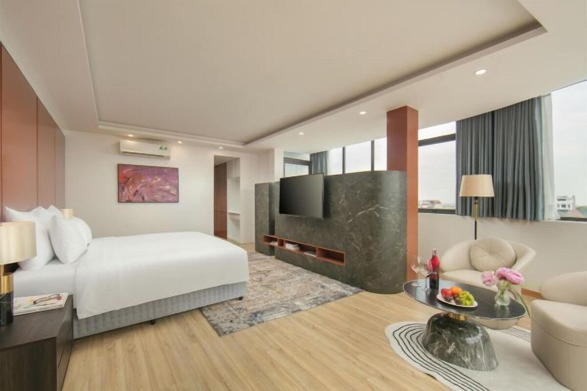 Une chambre d’hôtel moderne avec un grand lit, un téléviseur à écran plat fixé sur un mur de marbre, un canapé et une table basse agrémentée de fleurs.
