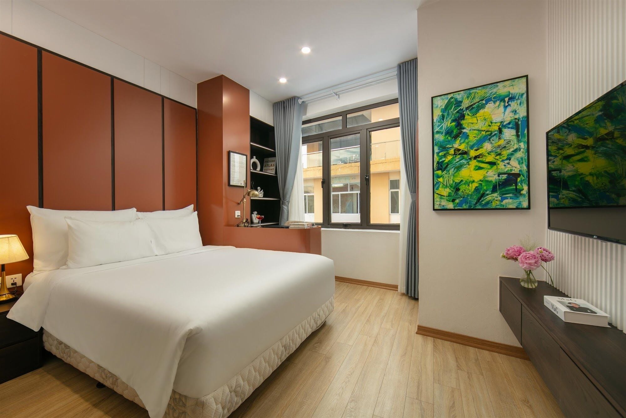 Hotelkamer met een bed, nachtkastje, bureau met lamp, raam met gordijnen en abstracte schilderijen aan de wanden.