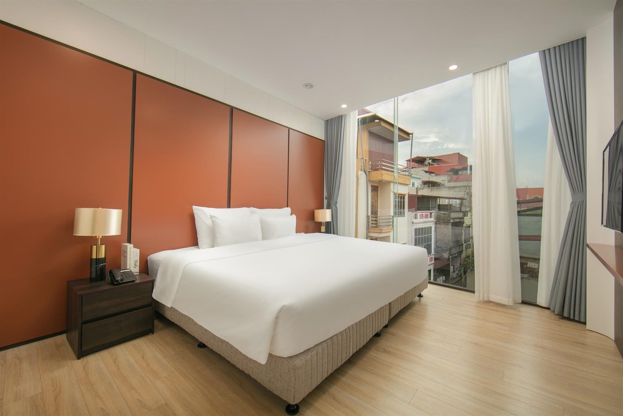 A hotel room with a large bed, a nightstand, a lamp, and a view of buildings through the window.