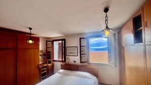 Un dormitorio con cama, armario de madera, escritorio con silla y ventana con vista a un tejado de tejas.