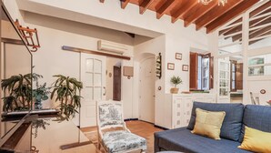 Un soggiorno con un divano blu, un tavolino in legno e una poltrona bianca con motivi floreali. Si nota anche un piccolo tavolino bianco con decorazioni a foglia, un pavimento in legno e un soffitto con travi in legno.