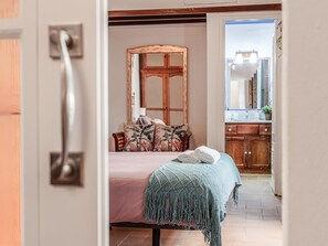 A bedroom with a bed, a wooden headboard, a bathroom with a mirror and sink, and a view of the outside.