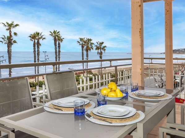 A dining table set with plates, cutlery, and glasses, overlooking a beach with palm trees and the ocean.