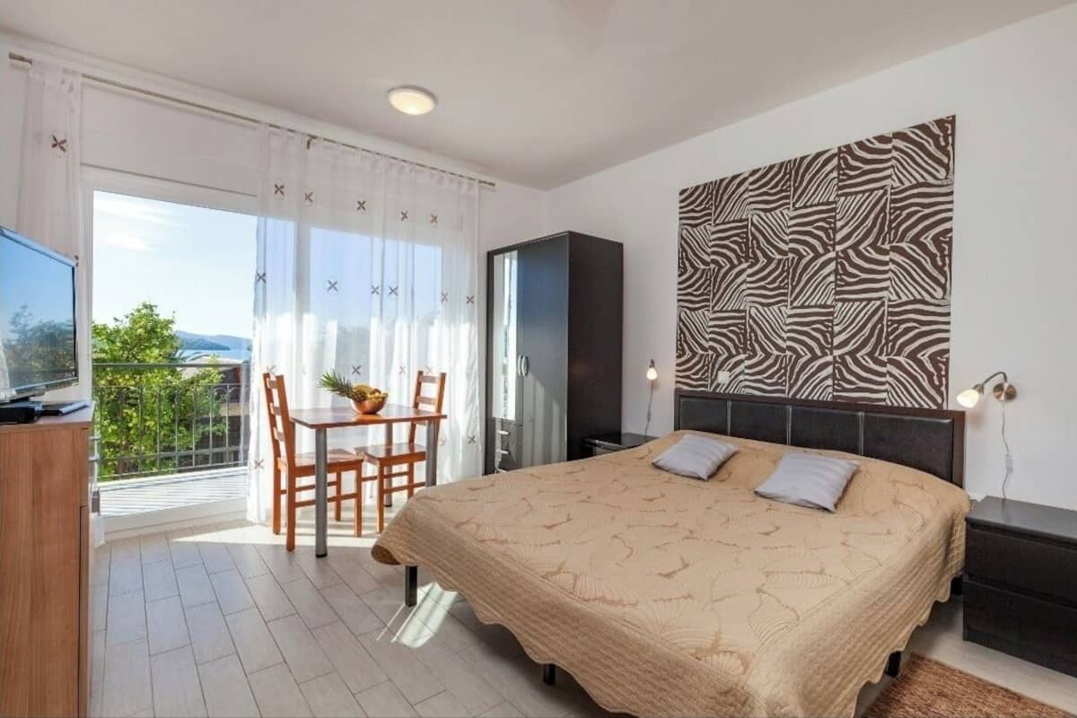 A bedroom with a large bed, a TV, a dining table with chairs, and a balcony with a view.