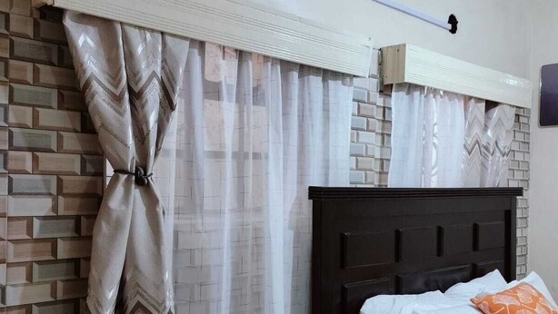 Un dormitorio con pared de ladrillos, una cama con ropa de cama blanca y un cojín naranja, y una ventana con cortinas translúcidas recogidas.