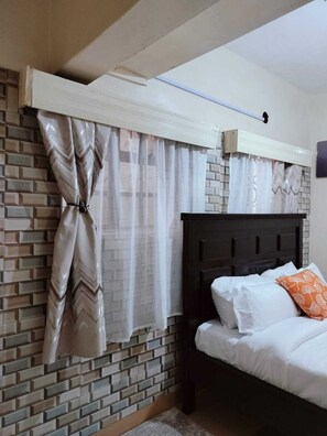 A bedroom with a brick-patterned wall, a bed with white bedding and an orange pillow, and a window with sheer curtains tied back. - Lux Suites Mancelona Flats Parklands (Nairobi)