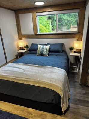 Un dormitorio con una cama grande, dos mesitas de noche con lámparas, una ventana con vistas a la vegetación y vigas de madera en el techo.