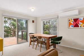 A room with a dining table, chairs, a sofa, a large window, and an air conditioning unit. - Island Treetops by Ready Set Stay (Smiths Beach)