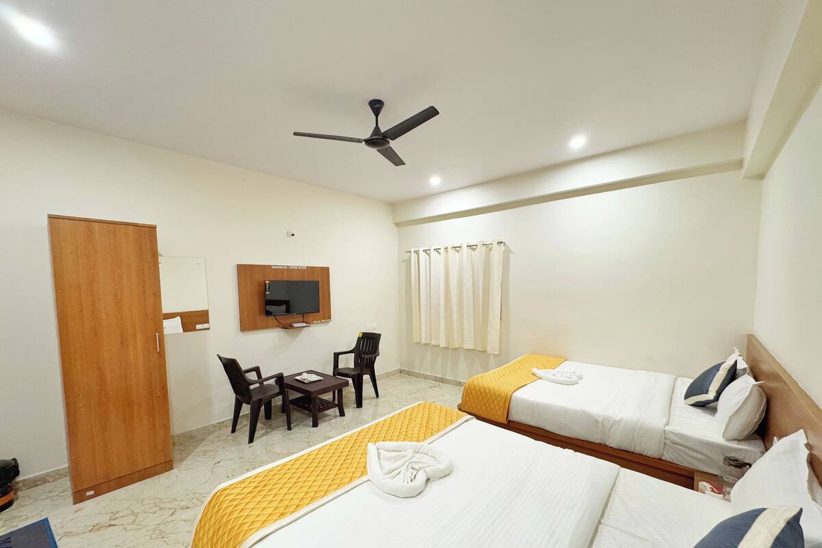 Ein Hotelzimmer mit zwei Betten, einem an der Wand montierten Fernseher, einem Deckenventilator, einem kleinen Tisch mit Stühlen sowie einem Badezimmer mit Vorhang.