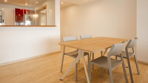 Un espace repas moderne avec une table en bois et des chaises grises, une cuisine visible à travers une cloison en verre, et un plafond blanc doté d’un éclairage encastré.