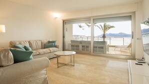 Un salon spacieux avec un canapé beige, une petite table ronde et une grande porte-fenêtre coulissante donnant sur un balcon extérieur avec vue sur la mer et les montagnes.