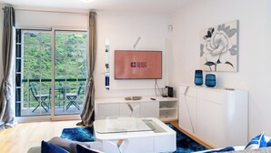 Un salon moderne doté d’une table basse en verre, d’un téléviseur à écran plat, d’une grande fenêtre offrant une vue sur la verdure et d’un décor minimaliste.