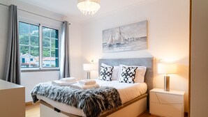 Une chambre moderne avec un grand lit, deux lampes de chevet, un tableau représentant des voiliers et une vue sur un quartier résidentiel par la fenêtre.