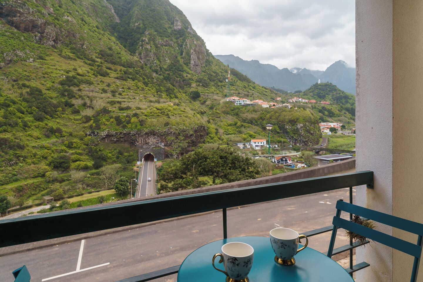 A balcony with two cups and a chair, overlooking a road with a tunnel and a mountainous landscape.