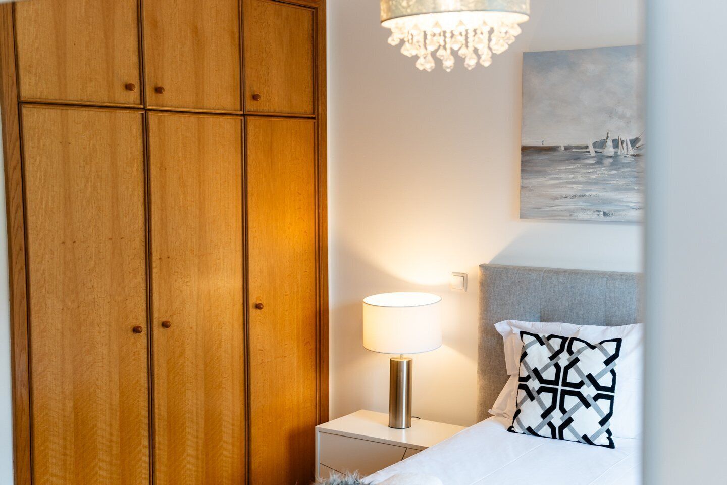 Un dormitorio con un armario de madera, una lámpara de noche, una cama con un cojín estampado y un cuadro en la pared.