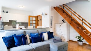 Un salon moderne avec un canapé d’angle gris, un escalier en bois et un coin cuisine doté de meubles blancs.