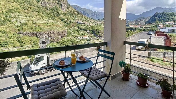 Un balcon avec une table et des chaises, donnant sur un paysage montagneux avec une route et des bâtiments visibles au loin.
