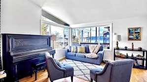 A living room with a piano, a blue sofa, two armchairs, a rug, and a large window with a view of a building and trees.