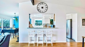 A modern kitchen with a large wall clock, white bar stools, and a view into the dining area.