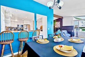 A dining area with a blue wall, wooden chairs, a table set for four, and a kitchen in the background.