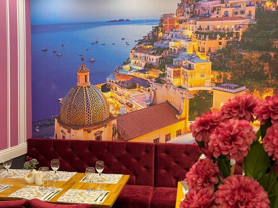 A restaurant with a mural of a coastal town, red velvet seating, and a vase of pink flowers.