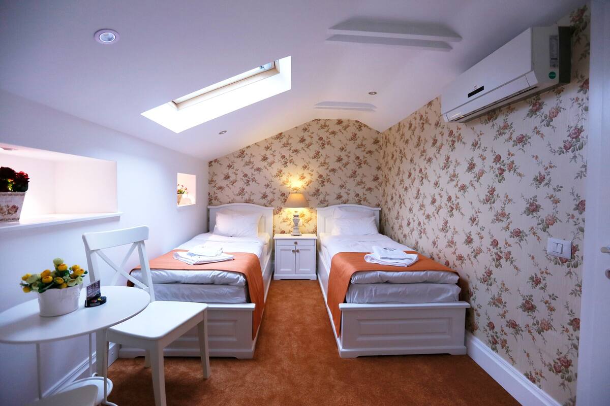 A small, well-lit room with two beds, a table, and a chair. There is a skylight above the beds, and an air conditioning unit on the wall.