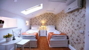 Una habitación pequeña y luminosa con dos camas, una mesa y una silla. Hay un tragaluz sobre las camas y un equipo de aire acondicionado en la pared.